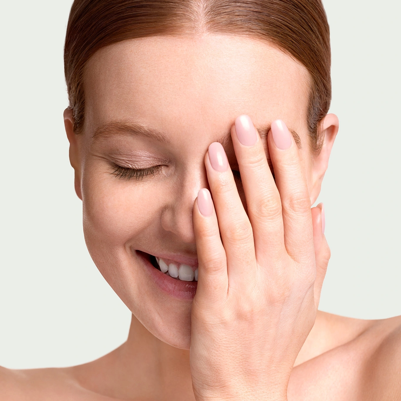 Woman smiling with hand covering face, eyes closed, fair skin, pink nails.
