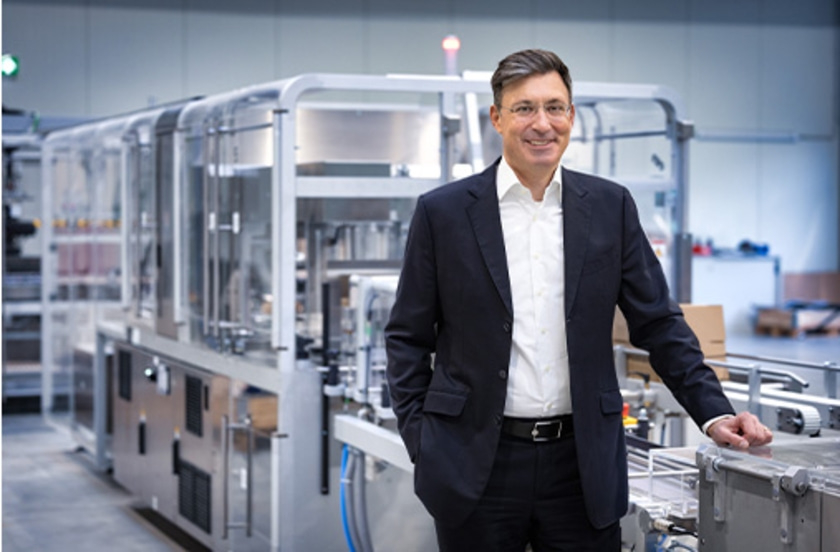 A man in a suit smiling in front of a machine in a factory.