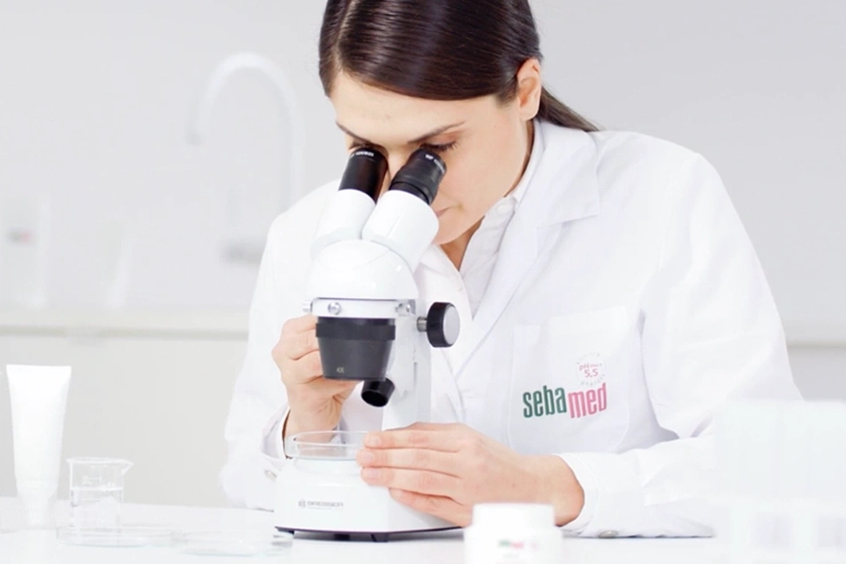 Científica mirando una muestra a través de un microscopio en un laboratorio, logo SebaMed
