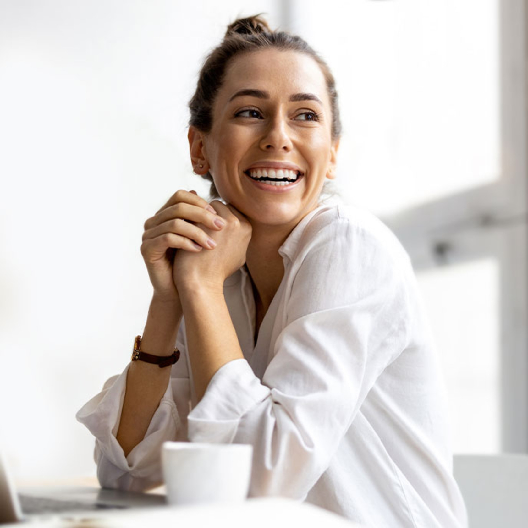 Mujer sonriente con las manos entrelazadas, camisa blanca, mirando hacia un lado.