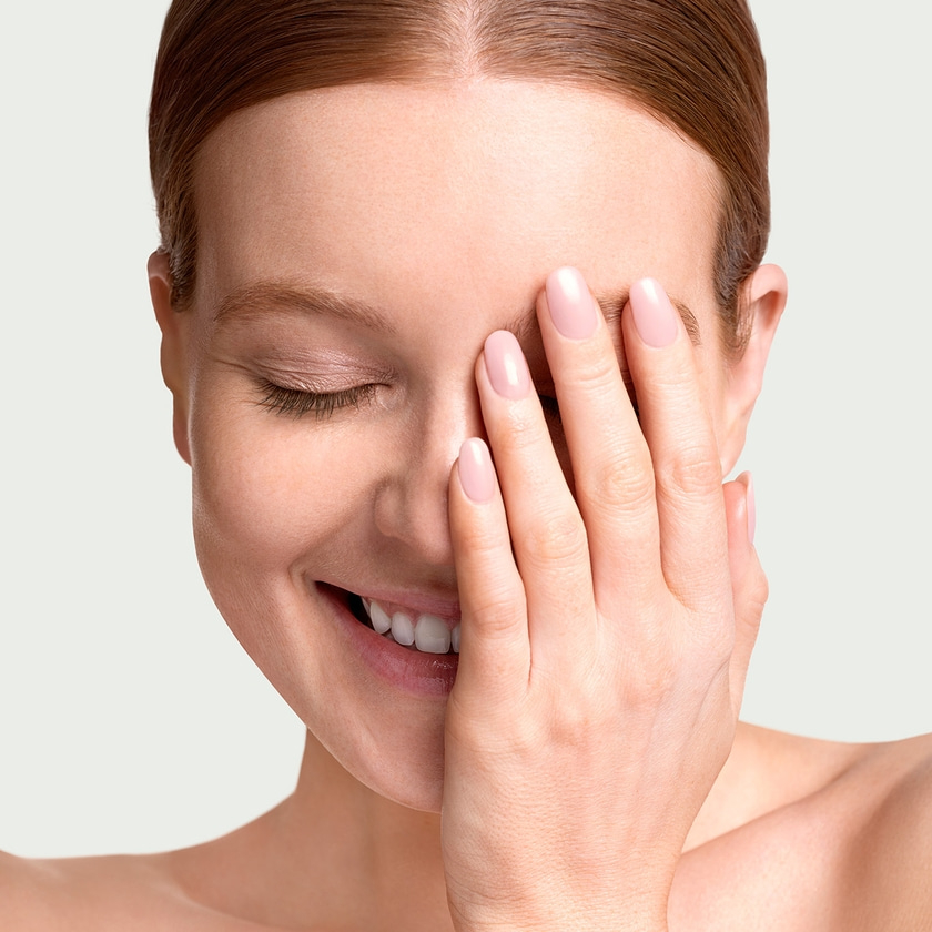 Woman smiling with hand covering face, eyes closed, fair skin, pink nails.