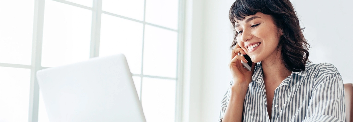 Woman smiling on phone, laptop, window, daylight, office, talking, happy, cheerful