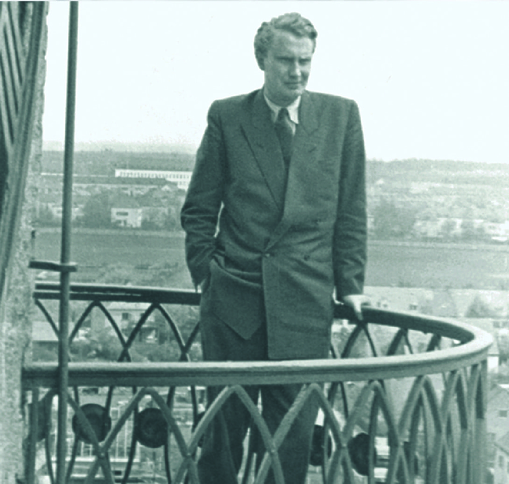 Man in suit on balcony overlooking landscape, hands in pockets, thoughtful.