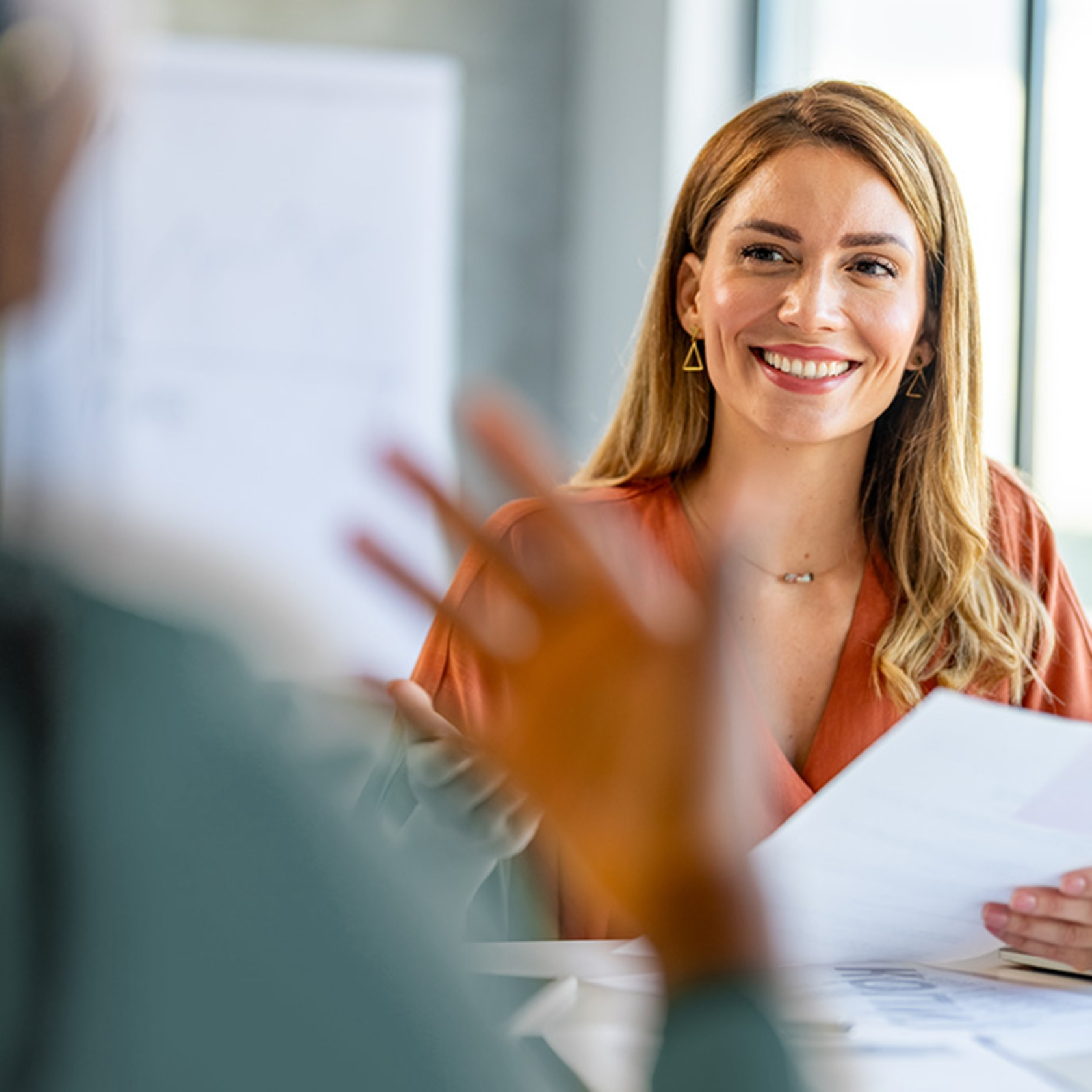 Smiling woman holding papers in an office, talking to someone.